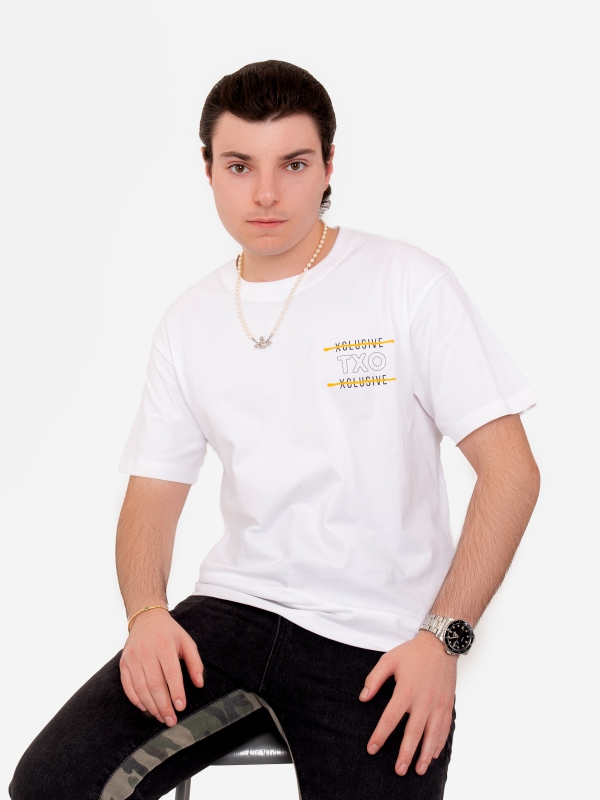 Young Man In A White Graphic T Shirt And Black Jeans Sitting On A Stool.